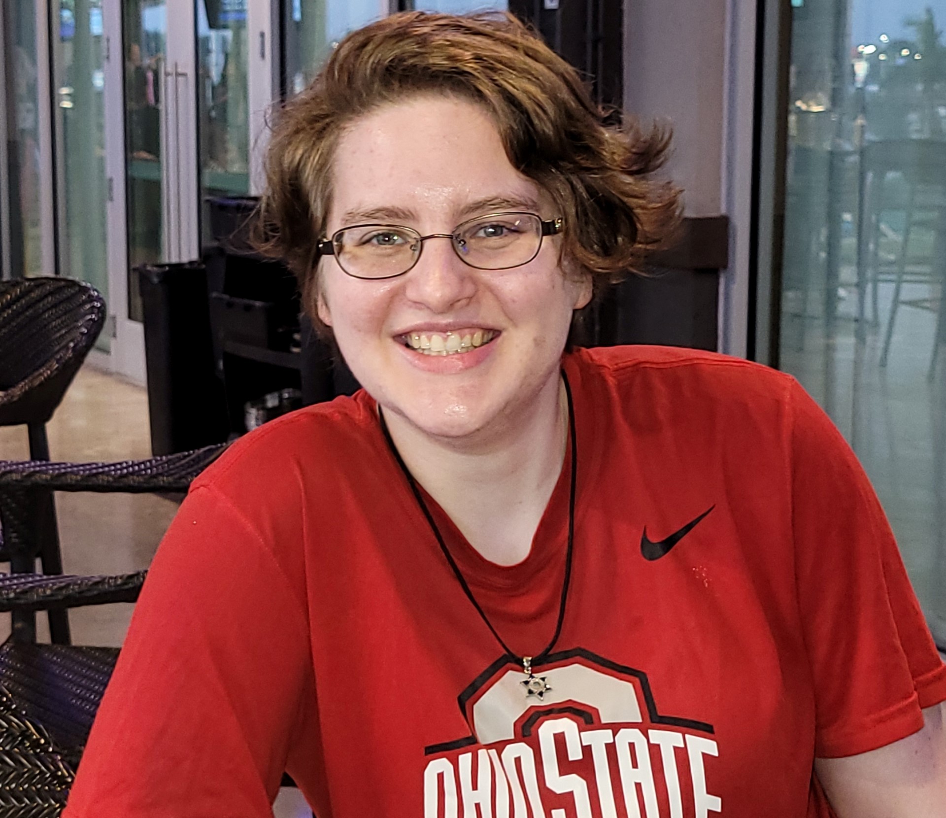 Photo of Saran Lendzian from the mid-torso up. They are wearing a red Ohio State shirt, a Star of David necklace, and glasses. They are white and have short, wavy, brown hair. They are posing informally and smiling. Photo of Saran Lendzian from the mid-torso up. They are wearing a red Ohio State shirt, a Star of David necklace, and glasses. They are white and have short, wavy, brown hair. They are posing informally and smiling.
